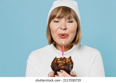 Close up elderly smiling satisfied cheerful caucasian woman 50s wearing white knitted sweater hold birthday cake blow candle isolated on plain blue color background studio. People lifestyle concept