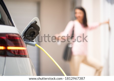 Close up of a electric car charger with female silhouette in the background, entering the home door and locking car