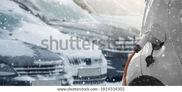 Close up of\
electric car with a connected charging cable on the blurred\
background of a snow covered parked\
cars.