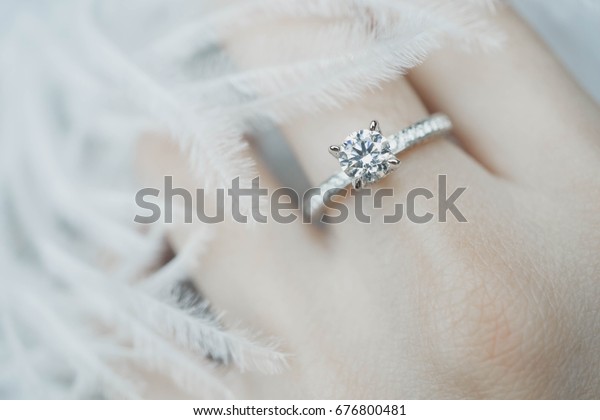 Close up of elegant\
diamond ring on the finger with feather and gray Scarf background.\
Diamond ring.
