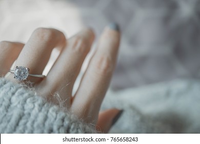 Close Up Of An Elegant Engagement Diamond Ring On Woman Finger With Dark Gray Sweater Winter Clothe Background. Love And Wedding Concept. Soft And Selective Focus.