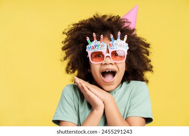 Close up of emotional curly little African American girl in party accessories while celebrating her birthday, isolated on yellow background. Party. celebration, fun, joy concept - Powered by Shutterstock