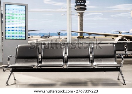 Close up of empty airport seating bench with plane runway and control tower visible on window. Airport waiting lounge with vacant metal chairs and electronic departure schedule display