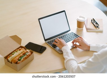 Close Up Of Entrepreneur Woman Hands Working On Laptop Computer Have Lunch Break Eating Sandwich On Desk At Home Office In Winter Day. Food Delivery, Quarantine, Take Out Food, Unhealthy Lifestyle.