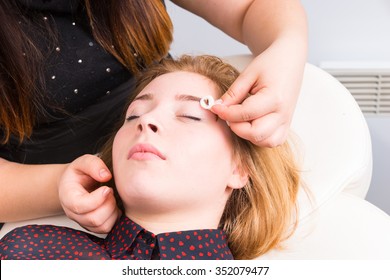 Close Up Of Esthetician Cleaning Up Excess Dye Coloring From Eyebrows Of Blond Female Client Reclined On Chair In White Spa, Using Cotton Pad