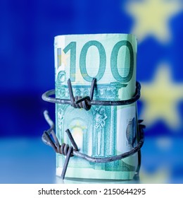 Close Up European Union Currency Wrapped In Barbed Wire Against Flag Of EU As Symbol Of Economic Warfare, Sanctions And Embargo Busting.