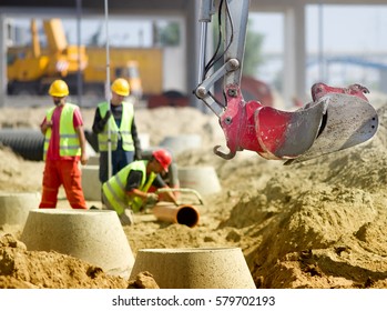 Close up of excavator bucket and workers working on manhole and sewerage pipes in background at construction site