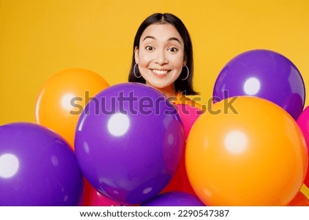 Close up excited surprised shocked impressed fun young woman wearing casual clothes celebrating behind bunch of balloons isolated on plain yellow wall background. Birthday 8 14 holiday party concept