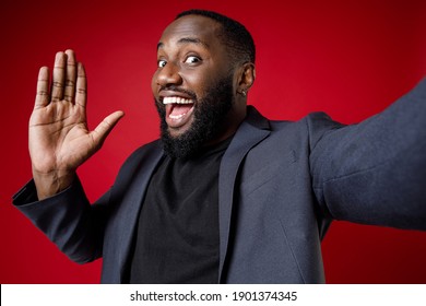 Close Up Of Excited Young African American Business Man In Classic Jacket Suit Standing Doing Selfie Shot On Mobile Phone Waving Greeting With Hand Isolated On Red Color Background Studio Portrait