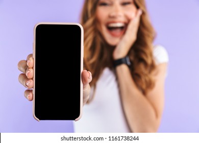 Close up of an excited young girl standing isolated over violet background, showing blank screen mobile phone