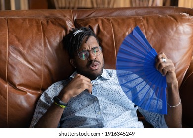 Close up exhausted African American man wearing glasses waving paper fan, cooling, suffering from overheating, hot summer weather without conditioner, relaxing on couch at home, breathing fresh air - Powered by Shutterstock