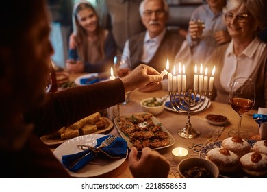 Close Up Of Extended Jewish Family Celebrating Hanukkah While Gathering At Dining Table. Focus Is On Father Lighting The Menorah.