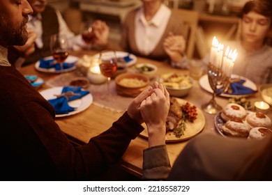 Close Up Of Extended Jewish Family Praying While Gathering Around Dining Table On Hanukkah. Focus Is On Couple Holding Hands.