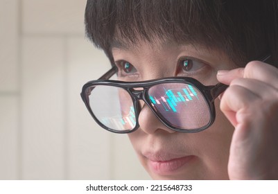 Close Up Face Of Asian Female Crypto Trader Looking At Computer Screen While Analyzing Online Trading Stock Market Data Chart In Office Room With Light Reflection On Eyeglasses Surface