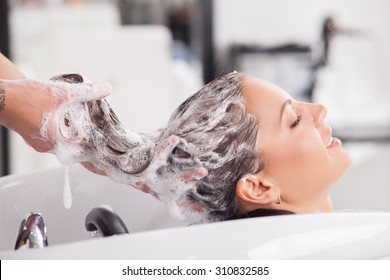 Close up of face of beautiful woman is getting a hairwash by a hairdresser. She is smiling. Her eyes are closed with relaxation. The beautician is making foam from shampoo - Powered by Shutterstock