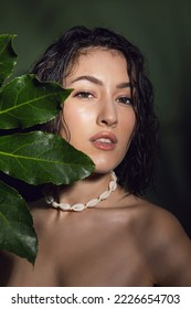 Close up face of beautiful young eastern woman covering face by green leaf on a background in the studio. Portrait of beauty woman with makeup and freckles, with wet hair