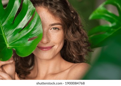 Close up face of beautiful young woman covering her face by green monstera leaf while looking at camera. Portrait of beauty woman with natural makeup and freckles standing behind big green leaves. - Shutterstock ID 1900084768