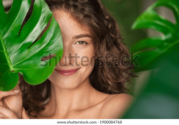 Close up face of beautiful young woman covering
her face by green monstera leaf while looking at camera. Portrait
of beauty woman with natural makeup and freckles standing behind
big green leaves.