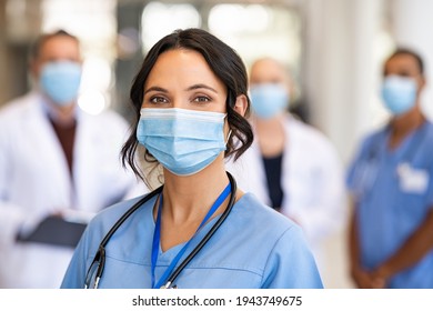 Close Up Face Of Confident Female Nurse In Front Of His Medical Staff Looking At Camera While Wearing Protective Face Mask Due To Covid-19 Virus. Smiling Surgeon Standing  With Team In Background.