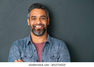 Close Up Face Of Handsome Mature Man With Beard Looking At Camera. Happy Mid Adult Indian Man Isolated Against Grey Wall. Portrait Of Smiling Mixed Race Guy On Gray Background With Copy Space.