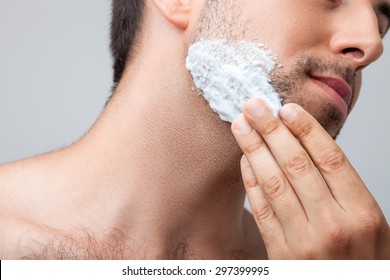 Close up of face of man applying foam on his chin with concentration. Isolated on grey background