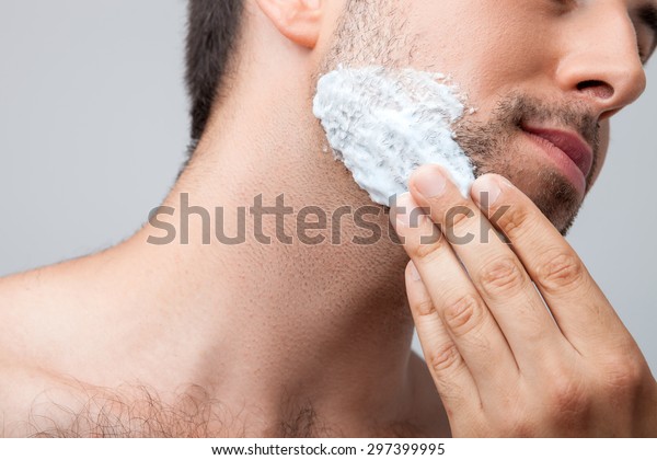 Close up of face of man\
applying foam on his chin with concentration. Isolated on grey\
background