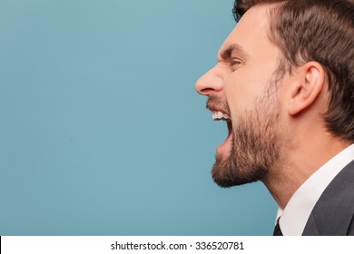 Close Up Of Face Of Man Shouting With Anger. He Is Standing In Profile. Isolated On Blue Background. Copy Space In Left Side