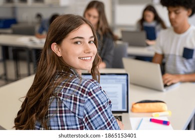 Close Up Face Of School Girl Looking At Camera While Studying On Computer. Portrait Of Smiling Young Woman Student Looking Behind While Using Laptop In University Library With Classmates In Background