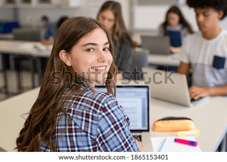 Close up face of school girl looking at camera while studying on computer. Portrait of smiling young woman student looking behind while using laptop in university library with classmates in background