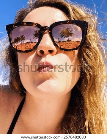A close up of a woman’s face. She has sandy, wind blown hair and is wearing reflective sunglasses. The reflection shows palm trees, a beach and a blue sky 