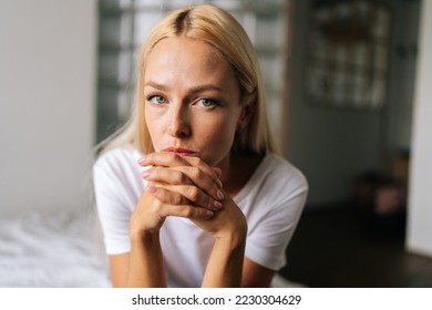 Close up face of thoughtful blonde female sitting alone in living room and serious looking at camera holding hands on chin, thinking over health problems, feeling sadness, boredom, apathy. - Powered by Shutterstock