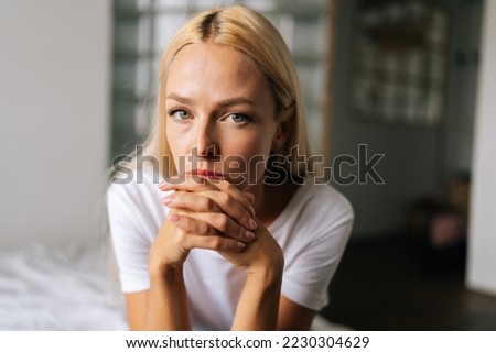 Close up face of thoughtful blonde female sitting alone in living room and serious looking at camera holding hands on chin, thinking over health problems, feeling sadness, boredom, apathy.