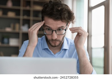 Close up face of young man in glasses stare wide-eyed at laptop screen, feel shocked by received news, confused or surprised by unexpected error, broken computer, overwhelmed with heap of scam message