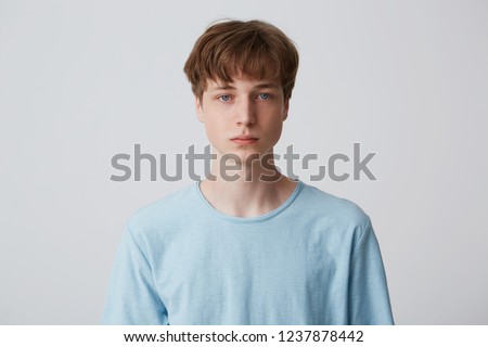 Close up face of a young man without emotions. Beautiful emotionless guy in a blue t-shirt looking to the camera, isolated over white background