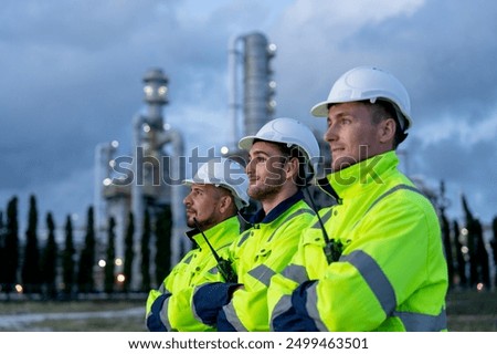 Close up factory engineer or technician workers stand in the row and look to left side with arm-crossed stand in front of refining and petrochemical factory during evening light. 