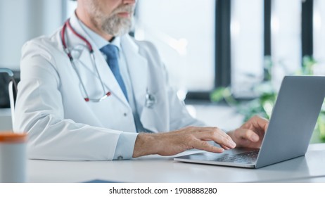 Close Up Of A Family Medical Doctor Working On A Laptop Computer In A Health Clinic. Physician In White Lab Coat Is Browsing Medical History Behind A Desk In Hospital Office.