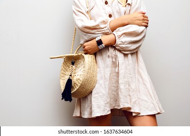 Nahaufnahme von Modedetails von Frau mit Leinenkleid, trendigem Strohhut und Accessoires, die auf cremeweißem Hintergrund posiert.