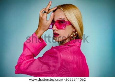 Close up fashion portrait of elegant woman wearing trendy pink sunglasses, fuchsia color blazrer, big golden earrings, ring, posing on blue background. Soft focus. Copy, empty space for text