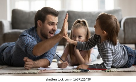 Close Up Father Giving High Five To Excited Little Kid Son After Finishing Drawing Picture. Smiling Dad Lying On Floor Carpet With Cute Children Siblings, Enjoying Creative Activity At Weekend Time.