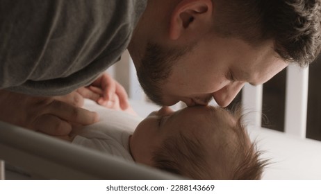 El acercamiento del padre juega y besa a un bebé adorable en la frente en una cuna infantil. Niño o niña vestido con traje corporal se acuesta en las sábanas de la mañana. Concepto de infancia, paternidad y familia. Movimiento lento.