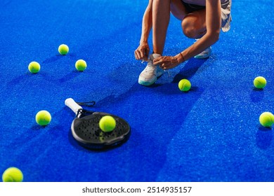 Close up of a female athlete tying a shoelace on her sneakers, kneeling down on a blue court next to a padel racket and padel balls. - Powered by Shutterstock