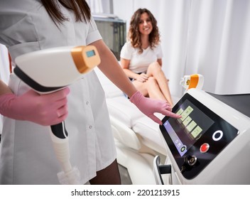 Close up of female beautician in sterile gloves using diode laser hair removal machine while smiling woman sitting on daybed. Esthetician preparing equipment for epilation procedure.