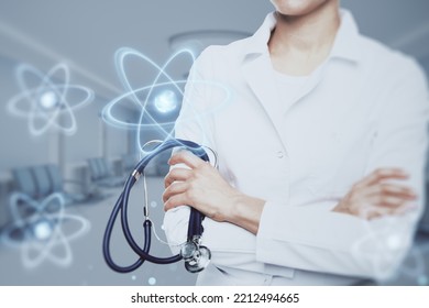 Close Up Of Female Doctor With Folded Amrs And Abstract Glowing Atom Hologram On Blurry Hospital Or Clinic Interior Background. Nuclear Medicine, Energy And Medical Research Concept