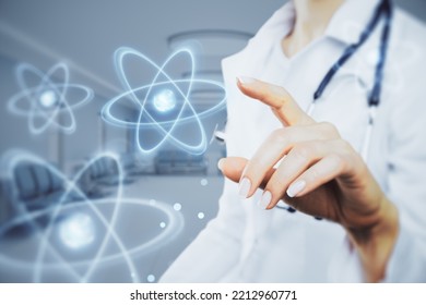 Close Up Of Female Doctor Hand Pointing At Abstract Glowing Atom Hologram On Blurry Hospital Or Clinic Interior Background. Nuclear Medicine, Energy And Medical Research Concept