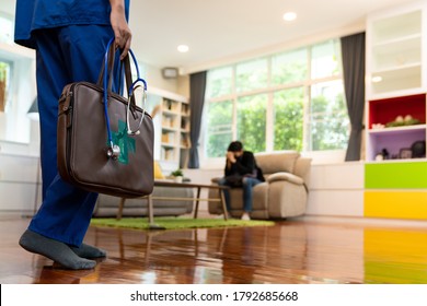 Close Up Female Doctor In Hospital Uniform With Background Of Asian Man With Face Mask Waiting In Living Room Of Apartment. Delivery Health Care Or Coronavirus Covid-19 Test At Home Concept.