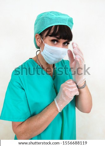 Close up of female doctor or nurse wearing face mask, stethoscope, hair net and green scrubs. Putting on clear latex gloves. Making eye contact. Plain pale background.