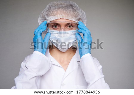 Close up of female doctor or scientist with a medical mask, surgical cap, glasses and hands in gloves over grey background.