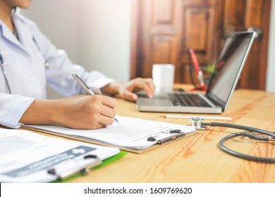 close up of female doctor writing notes on a paper on the clipboard, while using the touchpad on the laptop computer, representing medical research, working, and diagnosis of patient’s health care