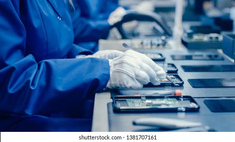 Close Up of a Female Electronics Factory Worker in Blue Work Coat and Protective Glasses Assembling Smartphones with Screwdriver. High Tech Factory Facility with more Employees in the Background. - Powered by Shutterstock