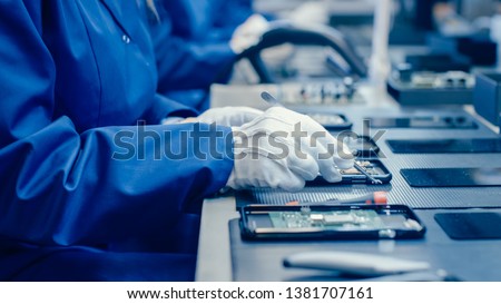 Close Up of a Female Electronics Factory Worker in Blue Work Coat and Protective Glasses Assembling Smartphones with Screwdriver. High Tech Factory Facility with more Employees in the Background.